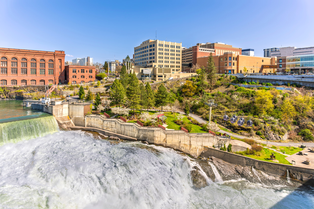 Spokane Falls cascading beside downtown Spokane, with riverfront paths, greenery, and city buildings overlooking the water