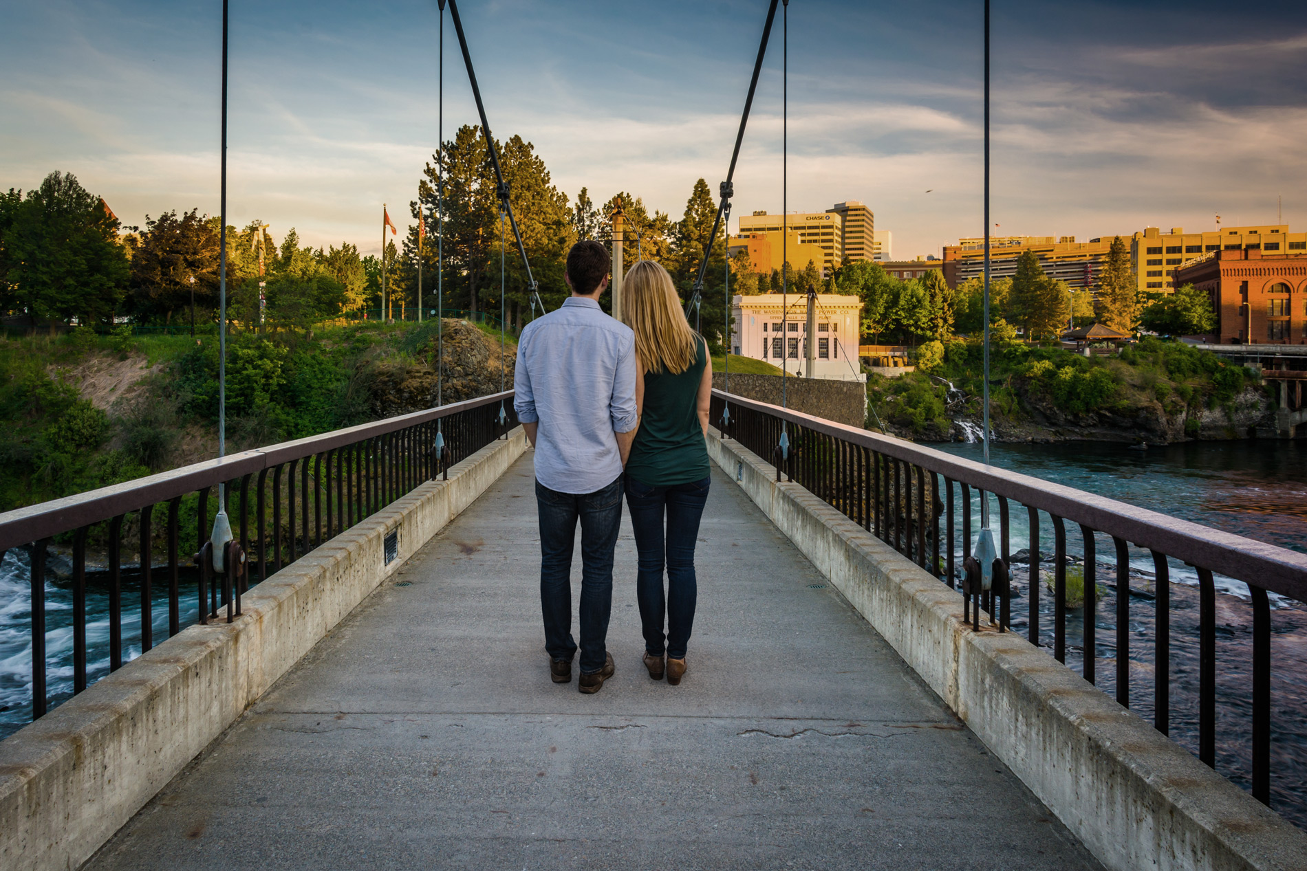 Couple standing together on a pedestrian bridge overlooking Spokane Falls, facing the city skyline at sunset