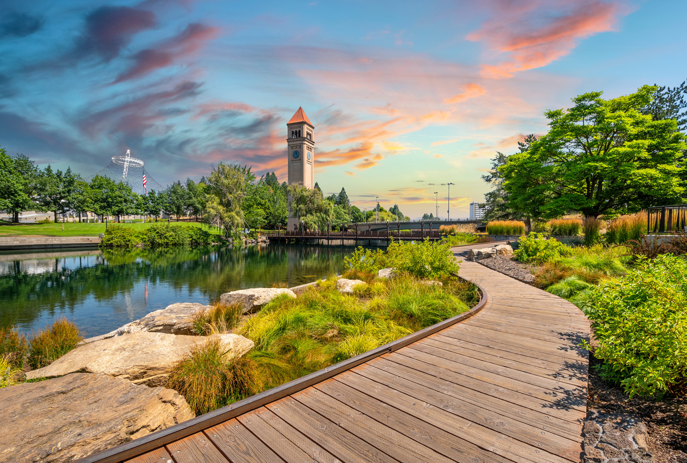 Riverfront Park in Spokane at sunset, with a wooden boardwalk path, landscaped greenery, reflective water, and the Clock Tower in the distance.